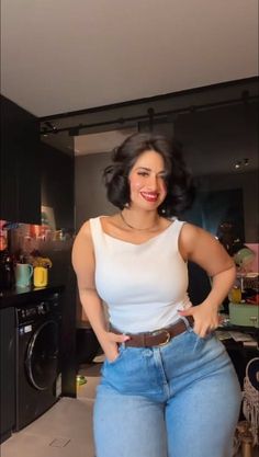Woman with short dark hair and red lipstick wearing a white tank top and jeans indoors