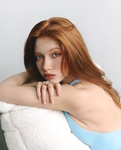 Woman with red hair and freckles wearing a light blue top, leaning on a white surface