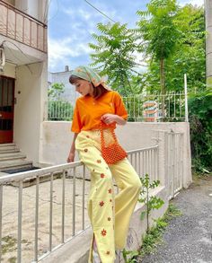 Woman in an orange t-shirt and yellow floral pants sitting on a fence outdoors