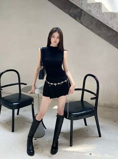 Woman in a black top and skirt with a chain belt and black boots indoors