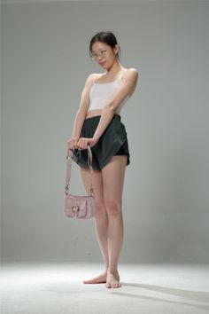 Woman in a white tank top and dark shorts holding a pink handbag in a studio