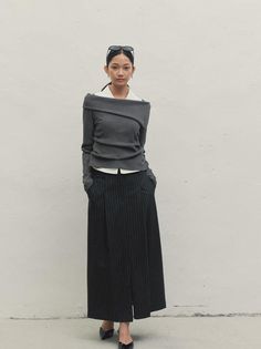 Woman in a gray off-shoulder top and pinstripe skirt standing against a white wall