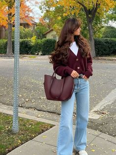 Woman with long brown hair wearing a cardigan and jeans on a street with autumn leaves