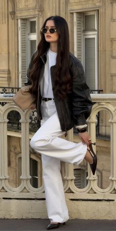 Woman with long hair and sunglasses wearing a leather jacket and white pants outdoors