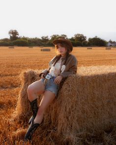 Woman in a cowboy hat and denim shorts leaning on a hay bale at golden hour