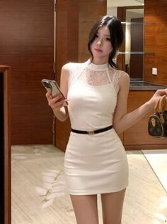 Woman in a white mini dress with lace detail holding a phone and handbag indoors