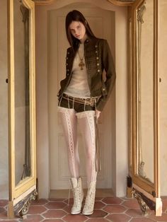 Woman in a military jacket, shorts, and white lace tights standing indoors