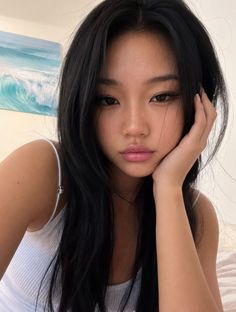 Woman with long black hair wearing a white tank top, hand on face