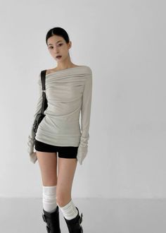Woman in an off-shoulder top, black shorts, and boots against a white wall