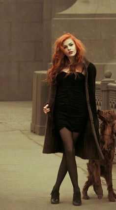 Woman with red hair in a black dress and long coat walking with a dog