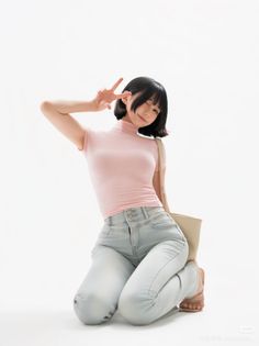 Woman with short black hair in a pink top and light wash jeans kneeling on a white background