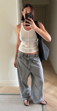 Woman in a white tank top and loose jeans taking a mirror selfie