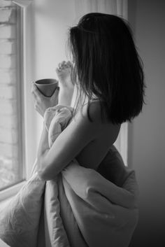 Woman wrapped in a blanket holding a cup, looking out a window