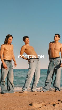 Three shirtless men in jeans on a beach with the ocean in the background