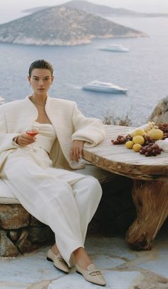 Woman in a white outfit holding a drink, sitting at a table with a sea view