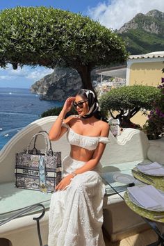 Woman in a white two-piece outfit and head scarf sitting outdoors with a sea view