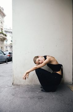 Woman in black top and pants squatting on a street next to a wall
