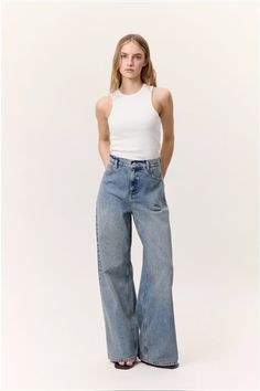 Woman in a white tank top and wide leg jeans posing in a studio