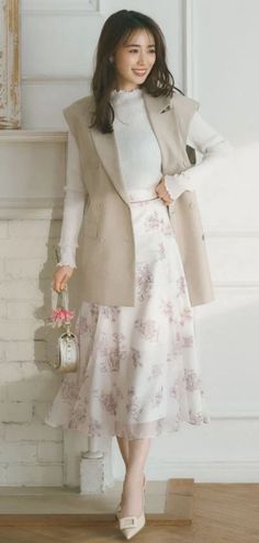 Woman in a floral skirt and sleeveless blazer holding a pink flower indoors