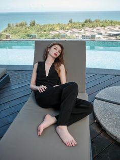 Woman in a black top and pants relaxing on a lounge chair by a swimming pool with an ocean view