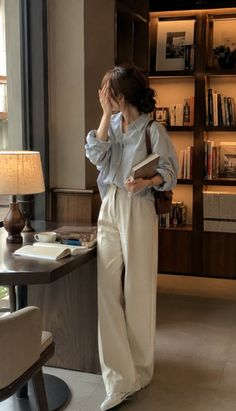 Woman in a light blue shirt and wide leg pants standing in a room with a bookcase