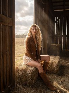 Woman with long blonde hair in a suede jacket and lace shorts sitting on hay bales
