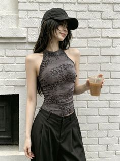 Woman in a leopard print top and black cap holding a drink against a white brick wall