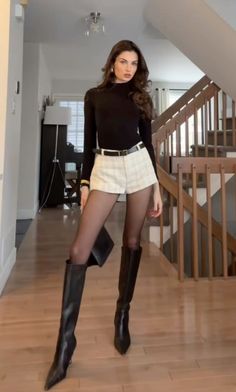 Woman in a black turtleneck, plaid shorts, and black boots standing indoors