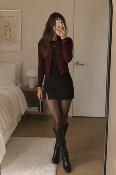 Woman in a black dress and cardigan taking a mirror selfie in a bedroom