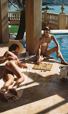 Two women in bikinis playing checkers by a swimming pool on a sunny day
