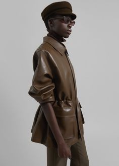 Person in a brown leather jacket, turtleneck, and newsboy cap with sunglasses
