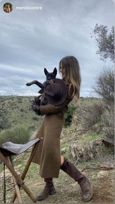 Woman in a brown dress and boots holding a dog and a hat in a hilly outdoor setting
