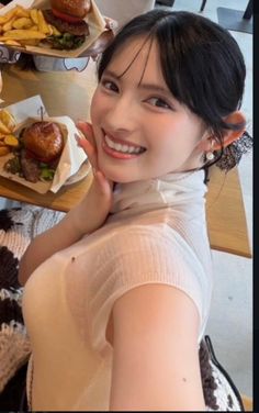 Woman with black hair wearing a white turtleneck, smiling at a table with food
