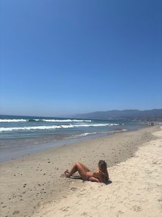Woman in a bikini lying on a sandy beach with ocean waves and mountains in the background