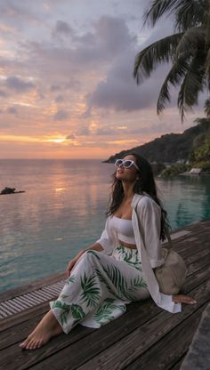 Woman in tropical print pants and white top sitting by an infinity pool at sunset