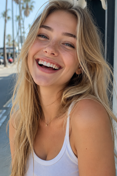 Blonde woman with freckles smiling broadly in a white tank top outdoors