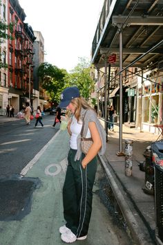 Woman in a baseball cap and track pants drinking a green beverage on a city street