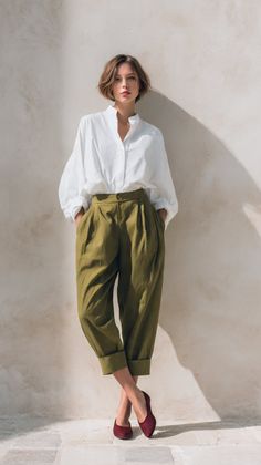 Woman with short hair in a white shirt and olive pants leaning against a wall