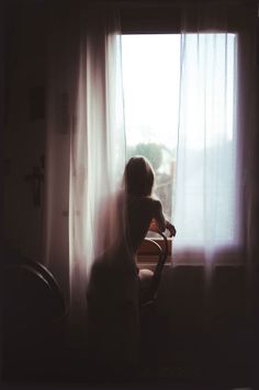 Woman with exposed back looking out a bright window from a dark room
