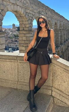 Woman in a black top and pleated skirt posing on a stone bridge with a cityscape in the background