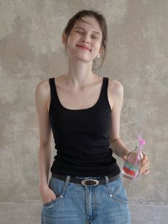 Woman in a black tank top and jeans holding a drink, smiling with eyes closed
