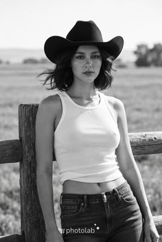 Woman in a cowboy hat and tank top leaning on a wooden fence in a field
