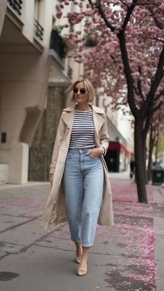 Woman in a trench coat and jeans walking on a city street with cherry blossoms