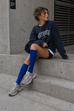 Woman with a messy bun wearing a blue sweatshirt and blue socks sitting on concrete stairs