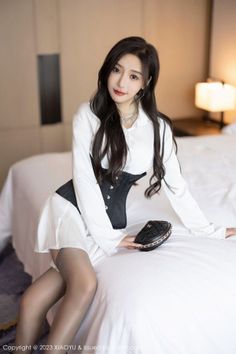 Woman with long black hair wearing a white shirt and black corset in a bedroom