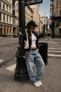 Woman in a black leather jacket and baggy jeans leaning on a lamppost in a city
