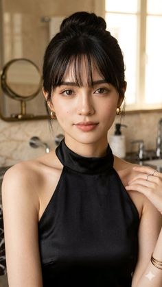 Woman with black hair in an updo and bangs wearing a black top and gold earrings