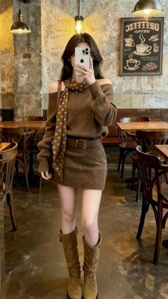 Woman in a brown sweater dress and boots taking a mirror selfie in a cafe