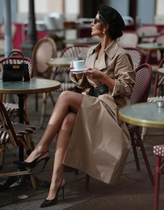 Woman in a trench coat and beret sitting at an outdoor cafe with exposed legs