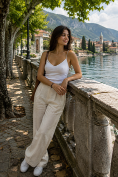 Woman in a white camisole and linen pants leaning on a stone railing by a lake
